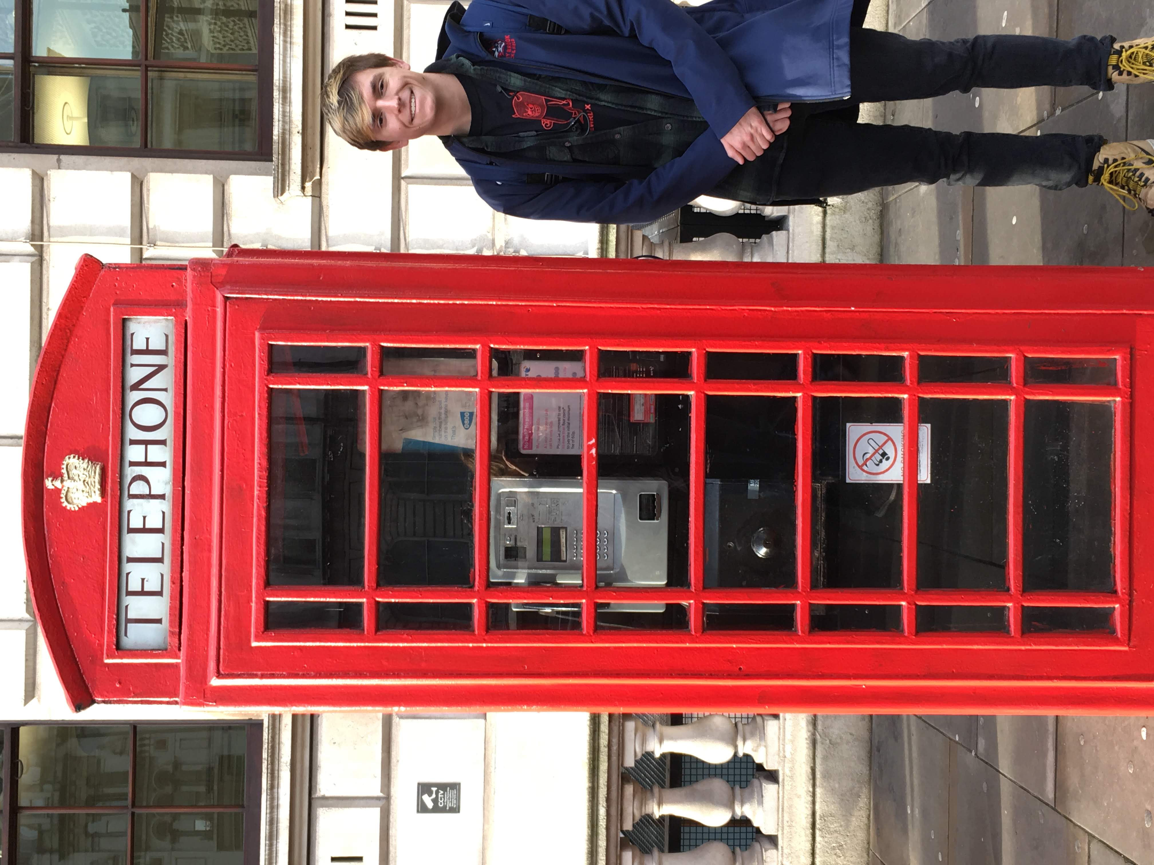 A young man with blonde hair and a smile stands on a sidewalk next to an iconic red British telephone booth. He is wearing a blue jacket over a black graphic t-shirt, dark pants, and tan boots. The background features a classic stone building with large windows and a decorative railing.