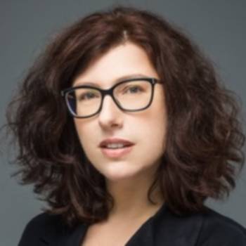 A professional headshot of a woman with voluminous, curly brown hair and glasses with dark frames. She has a neutral expression and is wearing a dark-colored blazer. The background is a solid, neutral grey.