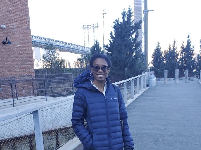 A smiling woman with short dark hair, wearing glasses, hoop earrings, and a navy blue puffer jacket, stands on an outdoor walkway. A red brick building is to her left, and a large suspension bridge is visible in the background under a clear sky.