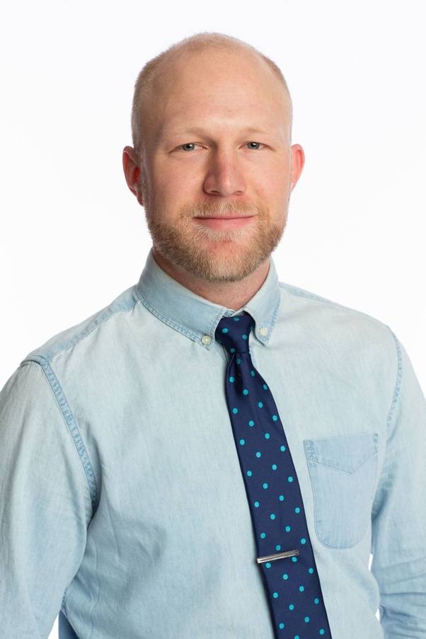 A professional headshot of a fair-skinned man with a shaved head and a light brown beard. He is smiling slightly and wearing a light blue denim button-down shirt paired with a navy blue necktie featuring light blue polka dots and a silver tie bar. The background is solid white.
