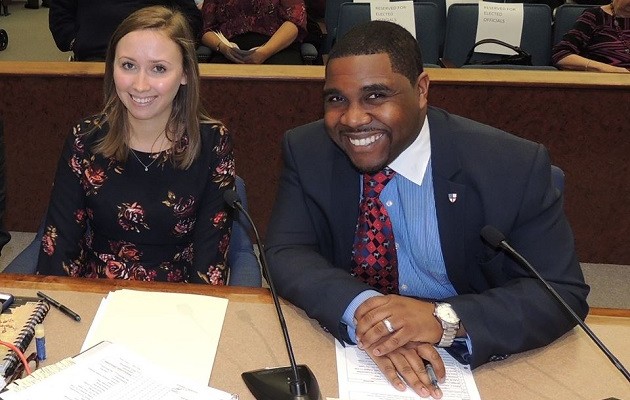 A smiling man and woman sit next to each other at a long wooden desk, likely in a courtroom or government chamber.  The woman on the left has long light-brown hair and is wearing a black long-sleeved dress with a red and pink floral pattern.  The man on the right is wearing a dark navy blazer over a light blue collared shirt and a red patterned tie. He has his hands clasped on the desk in front of him, showing a silver watch and a wedding band.  Two microphones are positioned on the desk, and various papers and a notebook are visible in the foreground. In the background, there are rows of blue upholstered seating.