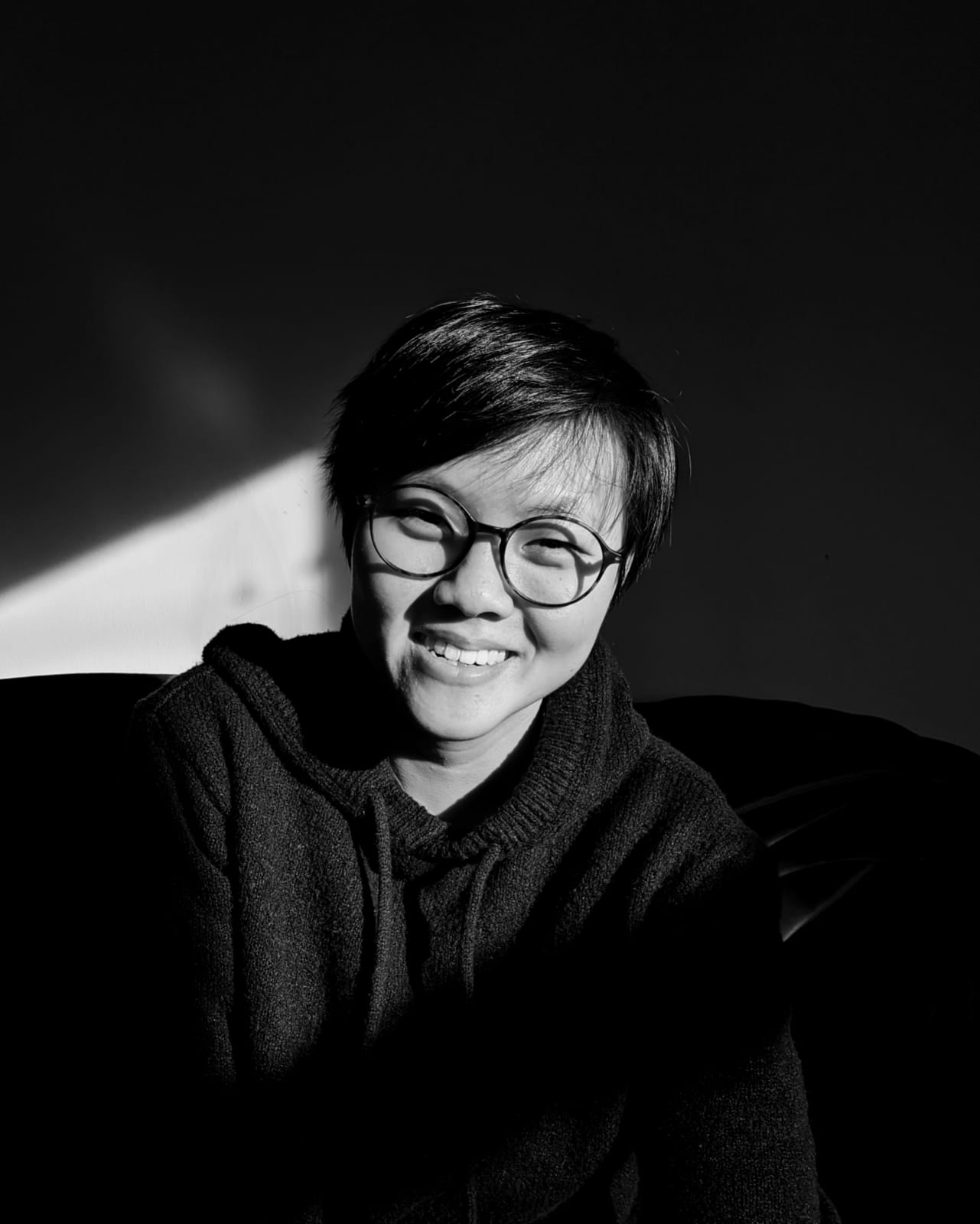 A black-and-white portrait of a smiling young person with short dark hair and glasses. They are wearing a dark hooded sweater. The image features high-contrast lighting, with a bright beam of sunlight striking their face and the wall behind them, while the rest of the scene remains in deep shadow.