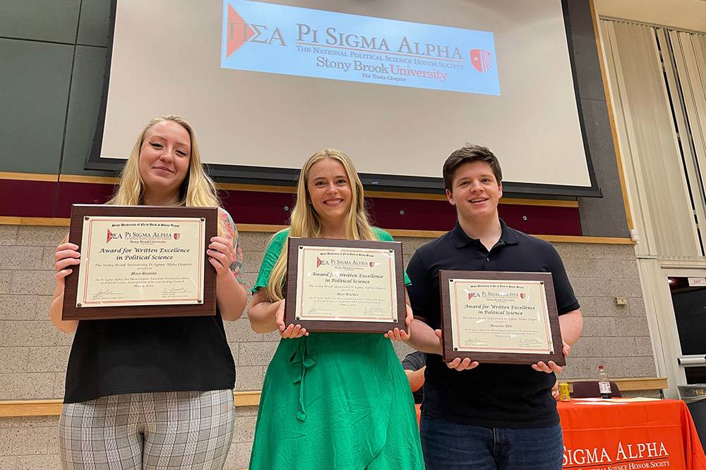 Three students—two women and one man—stand side-by-side on a stage holding wooden plaques that read "Award for Written Excellence in Political Science." They are posing in front of a projector screen displaying the Pi Sigma Alpha (The National Political Science Honor Society) logo for Stony Brook University. The woman on the left wears a black top and plaid pants, the woman in the center wears a bright green dress, and the man on the right wears a black polo shirt.