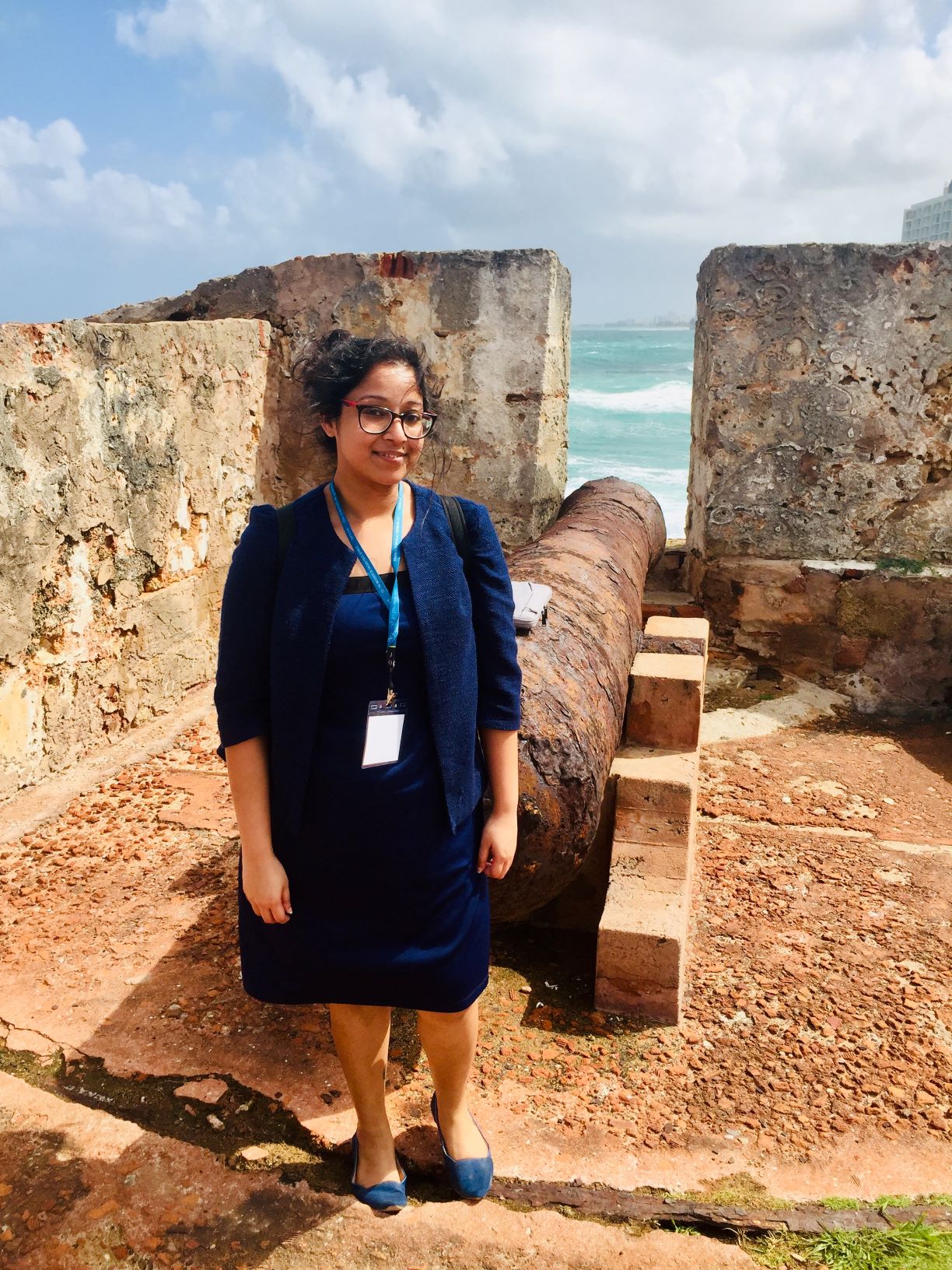A young woman with dark hair and glasses stands in an ancient stone fortification. She is wearing a navy blue dress, a matching cardigan, and a blue lanyard with an ID badge. Behind her is a large, weathered iron cannon pointing toward a gap in the stone wall, with the turquoise ocean and a cloudy sky visible in the background.