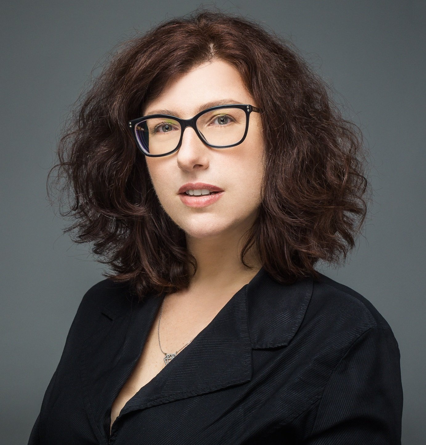 A portrait of a woman with voluminous, shoulder-length curly dark brown hair and black-rimmed glasses. She is wearing a black blazer and looking toward the camera with a neutral expression. The background is a solid, dark gray.