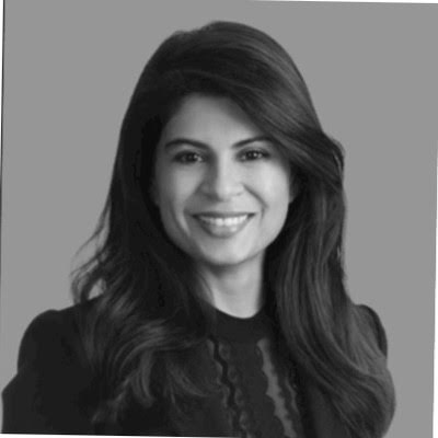 A black and white portrait of a woman with long, dark, wavy hair. She is smiling at the camera and wearing a dark, structured blazer over a top with subtle lace or embroidered detailing. The background is a solid, neutral gray.