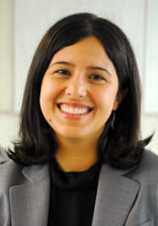 A professional headshot of a smiling woman with shoulder-length dark hair. She is wearing a grey blazer over a black top and small drop earrings. The background is a bright, neutral off-white color.
