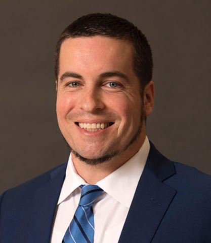 A professional headshot of a man with short, dark hair and a well-groomed beard and mustache. He is smiling warmly at the camera, wearing a white dress shirt, a blue-and-black striped tie, and a navy blue blazer. He is positioned against a solid, dark-brown background.