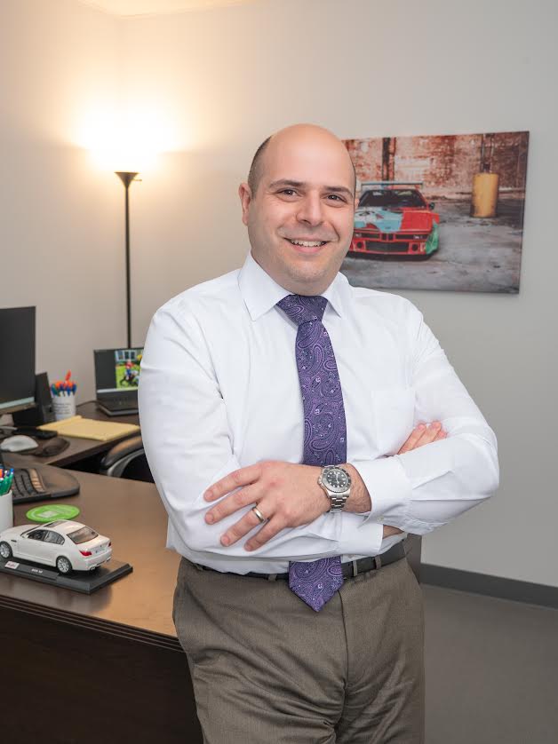 A professional portrait of a smiling, bald man with his arms crossed, standing in an office setting. He is wearing a white dress shirt, a purple patterned tie, and brown trousers. To his left is a dark wooden desk with a model car and office supplies, and behind him is a wall with a floor lamp and a framed picture of a red race car.