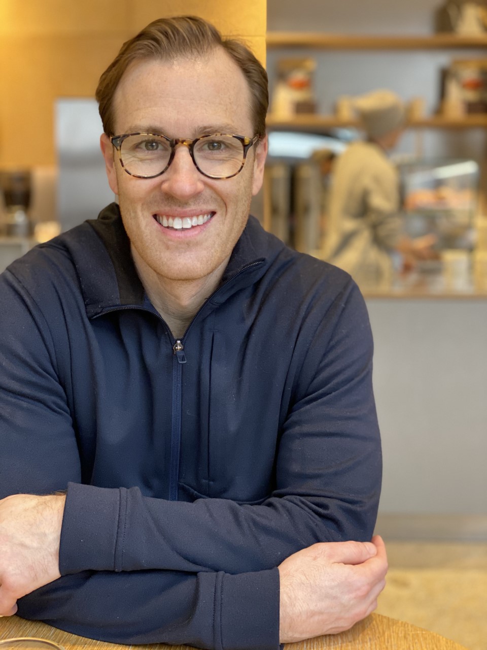 A portrait of a man with short brown hair and glasses, smiling at the camera. He is wearing a dark blue zip-up hoodie and leaning forward with his arms crossed. The background is a brightly lit, blurred indoor space that looks like a cafe or kitchen.